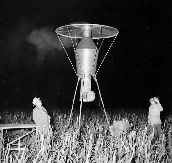 A vintage black-and-white photograph taken at night shows two men standing in a cornfield beside a tall, experimental frost-prevention machine. The metal device sits on tripod legs with a wide circular top and a conical center chamber emitting faint smoke into the dark sky. One man looks upward at the machine while the other stands near equipment on the ground, creating a dramatic scene of early agricultural technology testing.